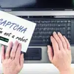 a pair of hands interacting with a laptop computer on a desk. The laptop is open, displaying a visible keyboard. One hand is holding a white paper sign with handwritten text that reads "CAPTCHA" along with a checkbox next to the phrase "I'm not a robot," which is checked. The paper also carries the logo "V6 Proxies".